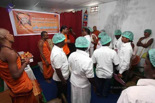 A relief-work room with a red curtain backdrop and a white banner reading Nithyananda Dhyanapeetam in English and Tamil, with a portrait of a saffron-robed figure. Several saffron-robed sannyasis with rudraksha malas stand on the left and centre, one holding a tray of apples. Around them, men in white shirts and white dhotis wear pale green hair caps, standing in a group and looking toward the front. A man in a white dhoti stands with arms folded at the far right. A blue floor mat covers the floor, and a white table with a red cloth is on the left.