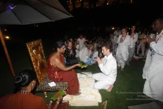 Outdoor evening gathering on a lawn at night under a large grey umbrella. A figure in a red-orange wrap sits on a leopard-print cushioned seat in front of an ornate gold-framed panel. Facing him, a young man in white kurta-style clothes kneels on a white fleece mat with palms pressed together, while a green leaf-wrapped offering is passed between them. Behind them, seated devotees in white and dark clothing, some with hands folded, and standing people in white with red beaded necklaces watch. A woman in an orange-brown garment at lower left holds a small wooden box. Lights glow in the background.