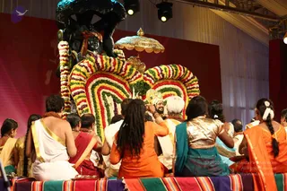 Photograph of a stage ceremony in front of a large black deity murti with a golden umbrella overhead and a raised arch of multicoloured marigold and rose garlands. Several people sit with their backs to the camera on a striped rug-covered dais in front. A man in white with gold-bordered shawl sits at left, women wear saris in orange, teal, red and gold blouses, and a person with long dreadlocks wears orange. A man at centre-right wearing a white shawl and ornate headwear faces the arch, and an older man in white sits beside him. Brass offering trays are placed on the dais.