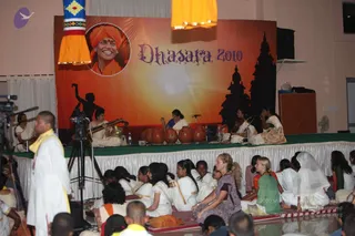 Indoor stage event at a hall during the Dhasara 2010 celebration. A large orange backdrop with a circular portrait of a smiling man in an orange turban and the words Dhasara 2010 sits behind musicians seated on white cloth on a green-edged stage. Several women in white saris play instruments: a stringed instrument, a row of large clay pots, and percussion. Silhouetted temple towers and a dancing figure appear on the backdrop. In the foreground, a man in white with a yellow shawl stands near a camera on a tripod, while many seated people in white and colourful saris and kurtas face the stage. Yellow lanterns hang from the top corners.
