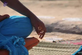 Close-up photograph of a seated person on a woven straw mat, shown from the side. The person wears a bright blue wrap over orange-brown cloth and has bare feet. The visible hand, with its fingers folded in a mudra, rests on the knee. The background is sandy ground with a patterned cloth on the right.