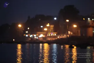 A blurred, long-exposure night view of a riverside ghat in Varanasi. Warm street lamps and illuminated shop fronts glow along the bank, their lights reflected in dark rippling water in vertical gold streaks. Silhouetted buildings and trees line the skyline under a deep blue night sky. No people are visible in the frame.