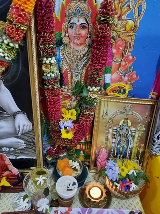 Home shrine photograph with no people visible. A large framed painted deity image in red and gold garments and jewelry, with a red and yellow flower garland draped across it, stands at centre. To the left, a black-and-white photograph shows a seated bare-legged figure with dark mala beads on the wrist and feet resting on a lotus-patterned surface. To the right, a gilt-framed print shows a blue-skinned standing deity murti with a tall crown in an arched temple gateway. On the table below sit clear glasses of water with jasmine flowers and leaves, a bowl of cut papaya, a cup of white offering, a lit oil lamp in a brass holder, and a bowl of mixed flowers including purple, yellow and pink blossoms.