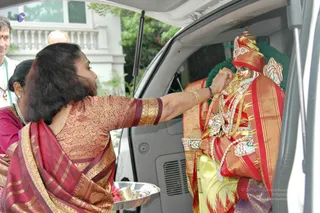 A woman in a red and gold silk sari with a gold-bordered blouse stands at the open rear door of a white vehicle, offering worship to a decorated deity murti seated inside. The murti wears a red and gold crown, a red and gold silk garment, layered white and silver jewelled necklaces and a green garland draped over the back. She holds a silver puja plate in her left hand and touches the murti's chest with her right hand, bangled and wearing a ring. Behind her, a man in a white shirt with a rudraksha-style mala and a woman in a maroon top stand partly visible at the left edge, with a white building and green foliage in the background.
