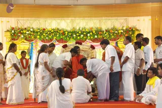 A ceremonial padapuja scene on a red-carpeted stage before a floral-decorated backdrop with gold-framed seating. A seated figure in saffron-orange robes sits on an ornate gold throne-like seat garlanded with red and white flowers. Several people in white dhotis and white saris kneel and bend before him, one washing or honouring his feet, while others in white stand around. A woman in a white sari with gold border stands at left and a girl in white with red-orange shawl stands nearby. Garlands of yellow, orange and red flowers hang along the top of the backdrop. The seated figure's appearance is not confidently established from the image, so he is described by role only.