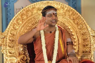 A man seated on an ornate gold throne with an elaborately carved gilded backrest, speaking and raising one hand in a gesture of explanation. He wears an orange-saffron cloth draped over his torso with a golden-yellow border, a white flower garland, a rudraksha mala necklace, beaded rudraksha bands on his upper arm, a red bindi and white sacred ash markings on his forehead, and a beaded headband. Behind him are blue and white painted walls. a person appears as the seated teacher, with a satsang discourse setting implied by the throne and microphone-wearing speaker.