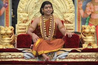 A man seated cross-legged on an ornate golden throne with red velvet cushions and golden lion-head armrests, draped over a tiger-striped cloth. He wears an orange-saffron garment with yellow trim, layered brown and gold beaded necklaces, gold armlets and bangles, a red bindi, and a small microphone at the ear. He gestures with both hands as he speaks. Behind him are large painted figures of deities with gold jewellery and red and orange backdrops. a person seated on a throne giving a discourse; the golden throne, saffron robes, rudraksha-style beaded mala and gold necklaces match the page's satsang context. One photograph with no other visible audience members.