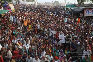 A dense outdoor crowd of many hundreds of people sits and stands on a dusty open ground at an event, many raising their hands and waving. Most wear white, cream, red, pink and yellow garments, with women in saris and shawls and some men in white kurtas. A few orange-clothed figures stand among them. Behind the crowd stands a row of tents, a green tarpaulin, a black canopy and a shop with a red and blue signboard. Orange flags are visible at the right edge. No identifiable central figure is clearly visible.
