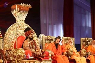 Photograph of a stage event with several seated men in saffron and orange robes on ornate golden thrones. On the left, a man in a red and orange turban, rudraksha-style beaded garland and gold armbands sits beside a golden cobra-hooded throne topped with a trident staff. Next to him, a bearded man in orange robes gestures with one hand. Further right, an elderly white-bearded man in orange sits on a gilded lion-armed chair, with another seated man at the far right edge. Golden lion sculptures and gilded framed portraits decorate the stage, with white drapes behind and a floor fan visible. The presenter on the far left is a seated man in ceremonial garb, and the scene suggests a satsang-style dais setting.