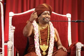 A man seated on an ornate silver-and-red throne in front of a red draped backdrop, speaking into a microphone on a stand. He wears a rust-brown turban with a gold-edged fold, a red tilak on his forehead, a rust-brown robe, a gold chain with a large gold pendant, and a long garland of white and pink flowers around his neck. He raises his right index finger while smiling, and a red thread is tied on his wrist. Swamiji (probable): long-established presentation cues of a turban, saffron-toned robe, ornate gold necklaces and rudraksha-style ceremonial dress, seated as the speaker of a discourse.