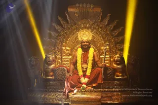 A seated man in a saffron-orange robe sits on an ornate gilded throne with lion-head armrests, leopard-print cushions and a tall golden hooded backrest. He wears a long yellow-and-red marigold garland, a crown-like gold headpiece, a gold necklace and bangles, and holds a staff in his right hand. Yellow spotlight beams light the dark stage behind him. The throne and lions are gold. Appearance-based label: a person, with long hair, saffron clothing and ceremonial gold adornments, seated in a dramatic stage setting.