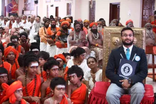 A large indoor hall filled with seated devotees and monastic participants in saffron and orange robes, many wearing orange turbans and white vibhuti forehead marks. In the foreground, many young boys with white painted foreheads wear orange cloth and rudraksha malas. On the right, a man in a dark suit, light blue tie and grey trousers holds a black folder labelled Guinness World Records Official and sits on a red cushion beside an ornate gold-embossed throne. Behind him, a bare-chested sadhu with a long beard and multiple beaded necklaces, and a figure with long hair and a crown-like headpiece, sit with hands folded. Wooden doors and framed pictures line the walls. The setting suggests a Guinness record presentation at an ashram.