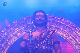 A man with eyes closed stands in front of an ornate golden arch lit in blue and magenta light. He holds a silver trident upright in his right hand and wears a leopard-print garment with rows of rudraksha-style beads across the chest, a gold necklace, and beaded bands on both upper arms. A vertical tilak mark is on his forehead, and his long dark hair is swept back. He is a sannyasi-style figure at a yoga session or darshan.