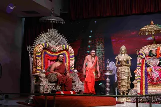 Stage photo of a ceremonial event with a seated man on an ornate silver throne draped in red, purple and yellow flower garlands, wearing a rust-red robe and a forehead tilak. Beside him a standing bare-chested man with long hair and a rudraksha mala wears an orange-red dhoti and folds his hands. To the right, a golden standing female statue with raised hands and a gilded deity murti with a canopy stand beside a brass kalasha-style lamp stand. Behind them a backdrop shows a glowing temple gopuram against a starry red sky. The seated man is an appearance-based candidate but the cues are insufficient to name him; the standing man has a rudraksha mala and a sparse ornate setup, so identity remains uncertain.
