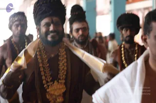 Photograph of a procession inside a temple corridor. In the foreground a smiling bearded man wears a dark maroon-brown turban with a vertical tilak on his forehead, layered gold ceremonial necklaces, and a brown robe draped with a white and gold-bordered shawl. Behind him, several men in similar brown robes, dark turbans or headwear and multiple gold beaded necklaces walk together, with a man in white dhoti and shirt at the right edge and other people blurred in the background among columns.