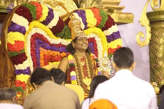 A seated figure on a richly decorated throne covered with garlands of red roses, yellow and white flowers, purple orchids and green leaves, with a golden crown and ornate gold necklaces. The figure wears a saffron-orange shawl and a rudraksha-style mala, holds a golden sun-shaped ceremonial emblem with a lion face, and smiles. Several people sit or stand in the foreground, seen from behind, including one in white and one in brown, with gold-pillared architecture behind. Swamiji (probable) is seated on the throne with a crown, saffron clothing and ceremonial gold necklaces, presiding over the satsang.