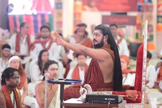 A photograph of a seated teacher in a rust-orange robe and matching shawl, with long dark braided hair and a full beard, raising his right hand in a gesture of instruction from a red velvet throne with silver carved armrests. Before him on a small blue-draped table sits a yamaha audio unit and a tray of incense sticks. Around him, a large seated audience in white ritual clothes and orange shawls listens attentively, with one woman in a white head covering and red-orange sari-style garments near the front. A young man in a white dhoti sits at left holding incense. Colourful patterned banners and an orange backdrop fill the hall behind.