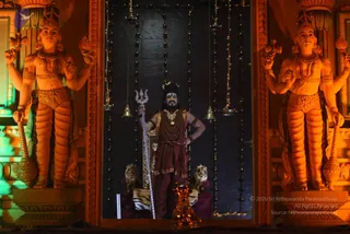 A figure stands in a lit niche framed by two large orange-painted guardian statues with multiple arms holding ritual objects. The central figure holds a silver trident in one hand, has a hand on the hip, and wears a maroon-red robe with a gold waist sash, a gold necklace and many beaded rudraksha malas; long dark hair is swept up. Strings of small lights and hanging brass bells frame the dark backdrop, with brass oil lamps and a lion-shaped pedestal beside him. The scene reads as a darshan setting; the page does not indicate that a person is present.