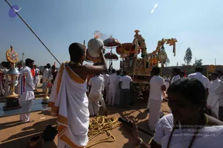 Outdoor ritual procession on dry earth under a clear sky. In the foreground a shaven-headed man in white dhoti with saffron-orange border raises a coconut-shaped kalasha vessel with a flame atop it. Behind him a decorated ratha-style cart with orange and gold canopies, garlands and a carved horse-like figure moves through a crowd of men in white dhotis and shirts. At left a tall silver and gold ceremonial urn-stand with colourful ribbons stands on a glass-topped table. A woman in white with a rudraksha mala holds a black tube-like object in the lower right. A coiled yellow rope lies on the ground. The page refers to Youth Day celebrations, but only the procession is visible.