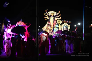 Night procession with a large illuminated deity murti of Devi on a decorated float, eight arms raised with weapons and a lion mount beneath her, flanked by a glowing ornate shrine. Participants in white and saffron-tinted clothes walk beside the float, many holding decorated umbrellas in purple, black and red; one figure on the left holds a trident. Colourful flags and a purple-lit stage area surround the scene. No a person candidate is clearly identifiable in the crowd. Several people are visible, including partially visible figures at the edges.