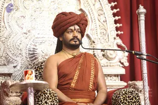 A seated man in an ornate silver throne with carved floral and scrolled patterns sits before a microphone on a stand, with a red curtain behind him. He wears a rust-red turban, a red-orange draped robe with gold-bordered edges, and a gold necklace, and has a red tilak mark on his forehead. Leopard-print cushions cover the throne arms. A small silver side table holds a framed photograph of a bearded man in orange. A silver staff stands at the right. The scene suggests a discourse setting.