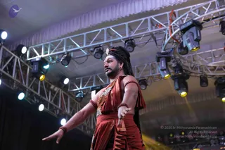 A performer on a stage under metal truss rigging and overhead stage lights, wearing a rust-red draped garment with a leopard-print patterned wrap, rudraksha-style bead bracelets on both arms, and a jewelled headpiece in long dark braided hair. He stands mid-motion with one arm extended outward and the other hand lifted, facing left in a dramatic dance-like pose. The background shows a dark curtain and a white ceiling drape. Only one person is visible. Based on the costume and presentation, this could be a person figure, though the appearance is not distinctive enough to confirm identity.