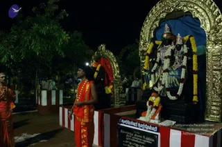 Night-time photo at a temple shrine with golden arched niche. On the right, two black stone deity murtis are adorned with yellow and white flower garlands and jewelled ornaments, with crowns on their heads, set on a blue backdrop. In front, a small seated figure with garland stands on a red-and-white striped peetha. A man in an orange-red dhoti and shawl with a gold-trimmed border and beaded necklace stands in the centre, hands folded. At left, a man in red robes with white forehead markings and folded palms stands beside him. Trees and a lamp-lit wall fill the background.