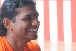 Close-up photo of a smiling man in profile, his dark hair swept back, with a red kumkum mark on his forehead and wearing an orange-saffron garment over the shoulders. Behind him are vertical bands of red and white. The orange robe and forehead mark suggest a sannyasi, but the face and appearance are not distinctive enough to confirm an identity, so the figure is described by role only.