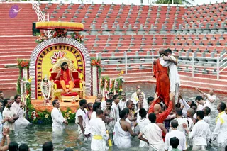 Outdoor ceremony at a red-tiered amphitheatre with a shallow water channel in front. A seated man on a gilded throne with a yellow canopy and flower-decorated arch wears saffron-orange garments and a rudraksha-style garland, and has long dark hair, consistent with a person appearance. Devotees in white dhotis wade in the water around the platform, some with raised hands and yellow sashes. In the water at right, a figure in orange robes embraces a man in white, while others lift hands toward them. A decorated deity murti stands at the left edge of the platform. Crowd of many devotees visible.