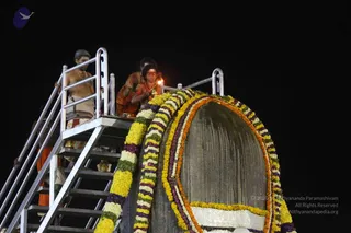 Night photograph of a large grey stone lingam decorated with bands of yellow, orange, white and purple marigold and flower garlands. On a metal staircase and platform beside it, a figure in an orange robe lights or holds a lamp with a visible flame, while a bare-chested man in white dhoti and gold chain stands at the rail on the left and another person in orange robes stands behind. Brass vessels sit on the platform. Dark sky background.