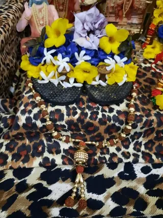 Close-up photo of a pair of dark, textured wooden padukas (sacred sandals) decorated with yellow allamanda and blue flowers, white jasmine blossoms and a lavender bloom, placed on a leopard-print cloth. A rudraksha mala with gold beads and a jeweled gold pendant with a tassel lies across the padukas. In the upper left, a partial statue wearing gold necklaces and a peach garment is visible, along with a red and yellow garland at the right edge. No people are visible.