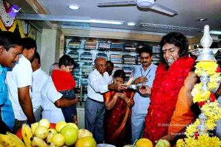 Photograph of a shop opening ceremony inside a retail showroom with shelves of folded shirts behind. A man in a saffron robe with long dark hair, a red bindi and a heavy red flower garland stands at right beside a silver aarti lamp decorated with yellow flowers, with a flame lit. Beside him, a woman in a maroon and gold sari holds a brass plate with a flame while an older man in white shirt and grey trousers assists. Other people stand nearby, one man holding a young child in a red top. Fruits including bananas, oranges and apples lie in the foreground, and garlands hang along the left edge. Swamiji (probable) is described by his saffron robe, long swept-back hair, red garland and bindi, and is presented as the guest of honour at this ceremony.
