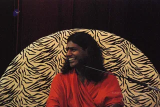 A smiling man with long dark hair sits in front of a large zebra-striped backdrop, wearing an orange-red shawl wrapped over his shoulders. A microphone stand arm is visible at right against the dark red curtain. The probable Swamiji is shown with long swept-back hair and saffron-orange wrap, seated and speaking, a discourse setting.