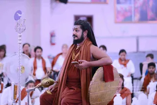 Photograph of a seated man in a rust-orange robe and matching shawl, with long dark dreadlocks, a full beard and a yellow-gold beaded necklace, seated on an ornate golden peetha-style chair with a red cushion. He holds a smartphone in one hand and a small golden bird-headed ritual object in the other. Behind him, a silver trishul stands beside a white fan. Rows of seated devotees in white clothes with orange shawls fill the hall, and framed artwork hangs on the wall.