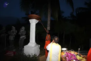 Pre-dawn outdoor scene at dusk-dark temple grounds with palm trees behind. A woman in an orange sari holds a long pole up to a large terracotta lamp bowl atop a white-wrapped pillar, lighting it. Beside her a man in white dhoti with a yellow shawl stands with his back to the camera, and a tray of flowers and brass items sits at lower right. Stone statues of standing deities stand at left beside a small red sign with partial text.