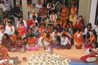 A large seated gathering of devotees on an orange cloth in an open hall, many wearing orange shawls or stoles. In the foreground a figure in orange robes seated at the left edge, with a microphone stand, beside a row of white plates holding bananas and sliced fruit with red offerings and dried stalks. Women in saris and salwar kameez, men in white kurtas and orange stoles, and children sit closely together, many with hands folded or attentive. A man in a dark jacket stands at the upper left. Several people wear rudraksha-style beads. The scene appears to be a satsang or puja sitting.
