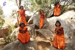 A photograph of five young girls in matching orange saffron-coloured saris with deep maroon borders, posed on large grey granite boulders outdoors among green trees. Several girls kneel or sit with palms pressed together in anjali mudra, one stands on a boulder with her arms raised, and another stretches one arm outward. Some wear rudraksha beaded necklaces and one wears a small headpiece. No a person candidate is visible, and the scene shows students in ceremonial dress posing together in a natural setting.
