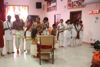 Indoor ashram hall with pink walls and tiled floor during a devotional line. A man with long swept-back dark hair and a red-brown shawl stands near the centre, in a line with several men and women in white dhotis and white kurtas, many wearing rudraksha malas and red sashes. A young girl in a white and gold outfit stands before an empty wooden chair with a red cloth draped over it, and a boy stands nearby. Participants have palms pressed together in anjali. Framed photos and a digital clock showing 08:20 PM hang in the background, alongside a large illuminated portrait. A plain bottle of water and a backpack sit at the edge of the frame.