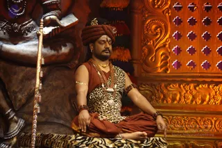 A man sits cross-legged with eyes closed in meditation on a tiger-striped seat in front of a large golden-brown statue of a deity with a raised trident-like staff. He wears a rust-red turban, a leopard-print sleeveless top, a rust-red dhoti, multiple rudraksha malas and a gold necklace, with arms adorned with rudraksha bracelets and a gold armlet. Behind him is an ornate golden carved wall with lattice panels and tassels.