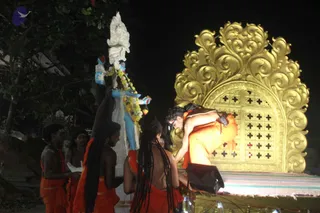 Night-time photograph of an outdoor ritual at a golden, ornately carved shrine-like structure with cross-patterned panels. A man in saffron-orange clothing bends forward over the platform, with a microphone and mic stand before him. Beside him stands a large blue-skinned deity murti with a white crown-like headdress and a garland of yellow flowers. To the left, several bare-chested men in orange cloth and beaded necklaces stand attentively, one holding a white cloth and another raising his arm toward the murti. Trees and darkness frame the scene.