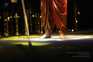 Close photograph of a standing person's bare feet on a dark stage floor, lit by warm yellow spotlight. The person wears flowing saffron-orange draped garments with a gold-bordered hem. A tall ornate golden staff with engraved patterns and a tassel stands beside the feet. Behind, rows of golden ceremonial lamps with lit flames line the dark background.