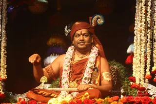 Photograph of a seated man on an ornate decorated seat framed by long garlands of white flowers and red roses. He wears a rust-orange head wrap with a peacock-feather style ornament, a bare chest draped in orange-red cloth, multiple rudraksha malas, a pink and white flower garland, and gold necklaces and armlets. He gestures with one raised hand while speaking into a headset microphone, with hanging tassels and lanterns behind him.