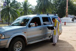 A silver Ford Endeavour SUV is parked on a sandy path beside tall coconut palms, with a white-painted small shrine and a tall decorated pillar behind it. A man in white dhoti and a yellow shawl stands beside the open rear door holding an open laptop, looking down at the screen. A person sits in the rear passenger seat and another figure is visible in the driver's seat. The vehicle bears a small blue-and-white logo sticker on its windshield. No appearance-based identification is made for any person in the scene.
