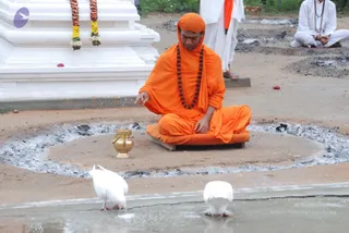 A photograph of an outdoor ritual at a temple-like setting. A figure in orange robes with a saffron head wrap and a long dark rudraksha mala sits cross-legged on an orange cushion inside a circle of grey ash, one hand raised and the other resting on the knee. A brass pot stands beside the cushion. White pillar with hanging marigold and yellow garlands behind. Two white doves drink from a shallow water channel in the foreground. Another person in white clothes with a rudraksha mala sits at the upper right, and a figure in white robes stands behind the ash circle. Small red flower petals lie on the ground.