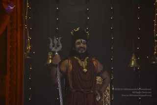 A photograph of a man standing in a dark, ornate hall lit by strings of small warm lights and hanging brass bells. He wears a maroon-red wrap and a long rudraksha mala, a heavy gold necklace, gold crescent-moon ornaments in his hair, and a white tripundra mark on his forehead. He holds a silver trident in his right hand and smiles, with gold-embroidered panels visible behind him. The presentation of a distinctive costume with crescent-moon crown, trident and rudraksha mala suggests a person in a stage-like Shiva-themed setting.
