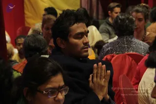A photo of a seated audience at a KAILASA satsang-style gathering in a hall with red and yellow drapery. A young man in the centre, wearing a dark jacket and a small tilak on his forehead, sits with eyes closed and palms pressed together in anjali mudra. Around him, devotees sit on red chairs and cushions, including a woman in the foreground wearing glasses and a bindi, and others in saffron, white and patterned clothing. No speaker or dais is clearly visible in this frame.