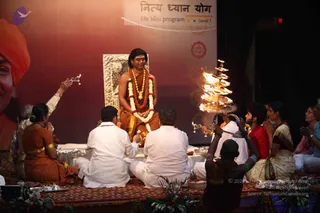 A seated figure in saffron-orange robes with a rudraksha-style necklace and a white-and-red garland sits on a carved silver-framed chair on a stage before a red backdrop. Several people sit on a patterned rug in white kurtas and saffron saris, facing him with hands folded or holding brass trays and small lamps. A tall tiered brass aarti lamp with flames is held up on the right, and flower arrangements and steel plates of offerings sit on the floor. A large portrait of a smiling man in an orange turban appears at the left edge. The stage banner reads a Hindi and English title on a life bliss program.