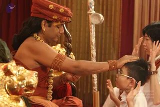 A seated figure in a red-orange turban with gold floral embroidery, wearing a rust-red shawl, gold beaded necklaces and rudraksha bracelets, touches the forehead of a young boy in glasses and a white shirt. The boy and a woman in a white top beside him have palms pressed together in devotion. Behind them hang gold-and-red drapes and a tall silver staff with a serpent-like wooden carving. A gilded lion-head armrest is at left. a person: the seated figure has long dark hair, gold necklace and rudraksha bracelets, and gives blessing to devotees.
