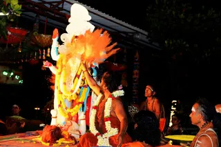 Night-time ritual photograph at an outdoor ashram setting with strings of paper lanterns and tassels overhead. At centre, a man with long dark hair and a bare torso, wearing an orange-red cloth and a white and red flower garland, raises his right hand toward a large white and gold figure of a deity murti, adorned with yellow marigold garlands and a white crown; a burst of orange powder or flame-like colour rises beside it. In the foreground, orange-clad devotees sit on the ground and watch, one with a white-painted forehead. Behind, a bare-chested man with white forehead markings and beaded necklaces stands, and a seated person on the right wears white tilak lines and rudraksha beads. Offerings and marigolds lie on an orange cloth at lower left.