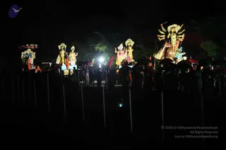 A night procession photographed from a distance, with several illuminated deity figures on floats or carts advancing in darkness. The largest figure on the right is a multi-armed goddess with a crown and red-and-yellow garment, standing on a red-draped base beside a lion-like form. Adjacent floats carry figures in gold and white crowns, with lamps and a small pale-coloured figure on the left under a striped canopy. Below them, a crowd walks with dark umbrellas held overhead, some people in white and saffron-toned clothing, with dim lighting and barricade posts in the foreground. The scene conveys a festive deity procession.